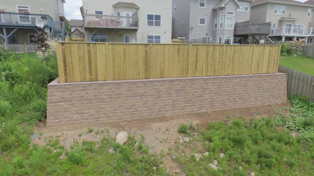 A wooden fence atop a stone retaining wall by Brent's Landscaping, Dartmouth's best. Lush grass and plants thrive below, with charming houses in the background under a partly cloudy sky. - Brent's Landscaping