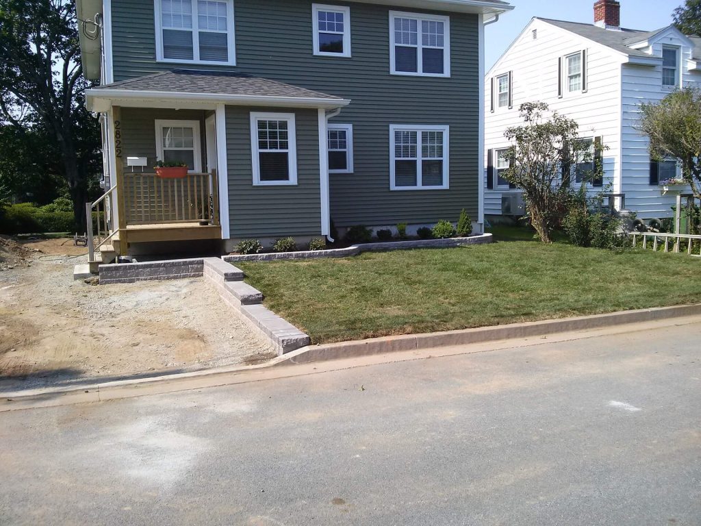 A two-story green house with a porch and steps leads to a front yard expertly landscaped by Brent's Landscaping, Dartmouth's best. Fresh sod, mulch, and shrubs create beautiful borders, with a white neighboring house visible on the right. - Brent's Landscaping