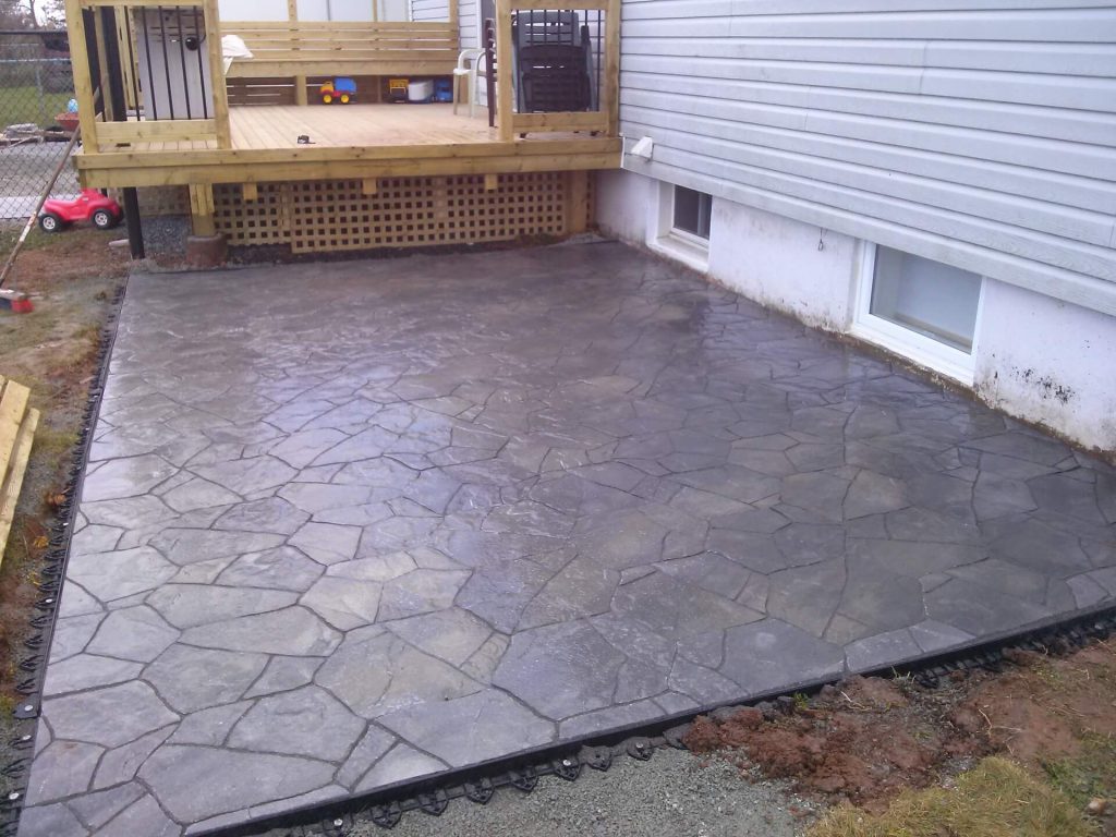 A stunning gray concrete patio with a flagstone pattern beside a white house, topped by a wooden deck with lattice panel, expertly crafted by Brent's Landscaping, Dartmouth's best. - Brent's Landscaping