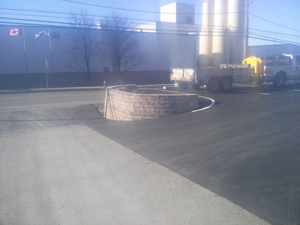 A newly constructed roundabout with a raised circular brick wall showcases Brent's Landscaping, Dartmouth's best. Freshly paved asphalt surrounds the area, and nearby are parked trucks, construction equipment, and fluttering flags. - Brent's Landscaping