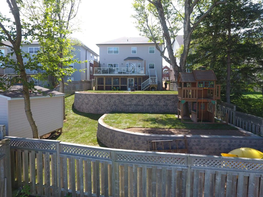 A charming backyard with a two-story gray house and elevated deck overlooks a stone terraced garden crafted by Brent's Landscaping, Dartmouth's best. Playground framed by fence and trees completes the idyllic scene. - Brent's Landscaping