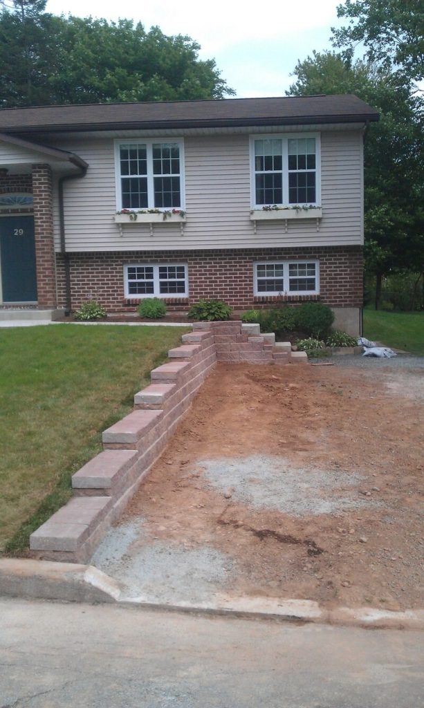 Image of a brick retaining wall and partially constructed driveway by Brent's Landscaping, Dartmouth's finest. House with brick/siding, two windows, amidst lush grass and trees. - Brent's Landscaping