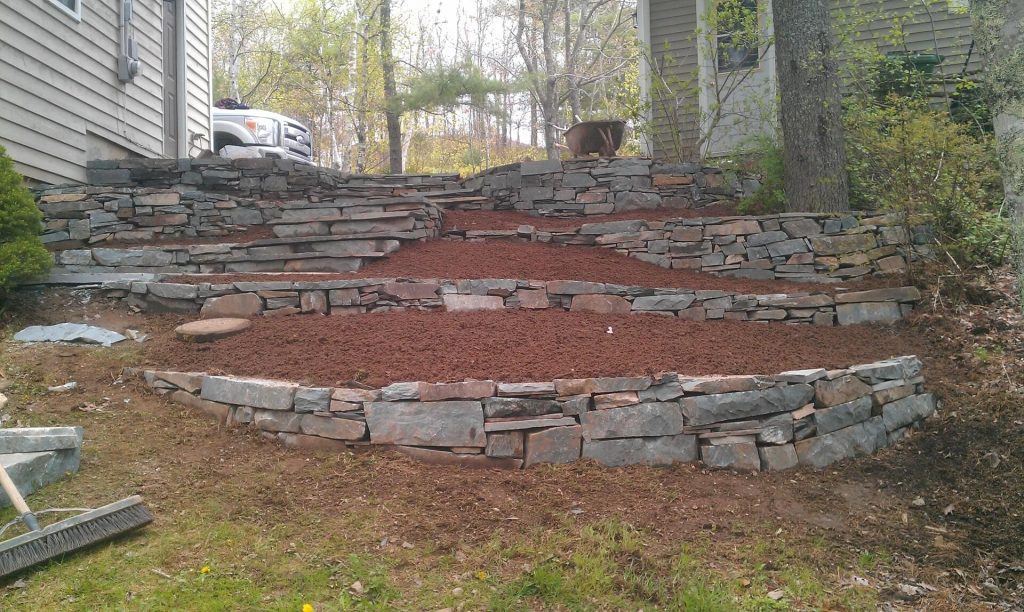 A landscaped garden with terraced soil levels, crafted by Brent's Landscaping, Dartmouth's best, is enclosed by stone walls and surrounded by trees and houses. A wheelbarrow rests on the upper terrace. - Brent's Landscaping