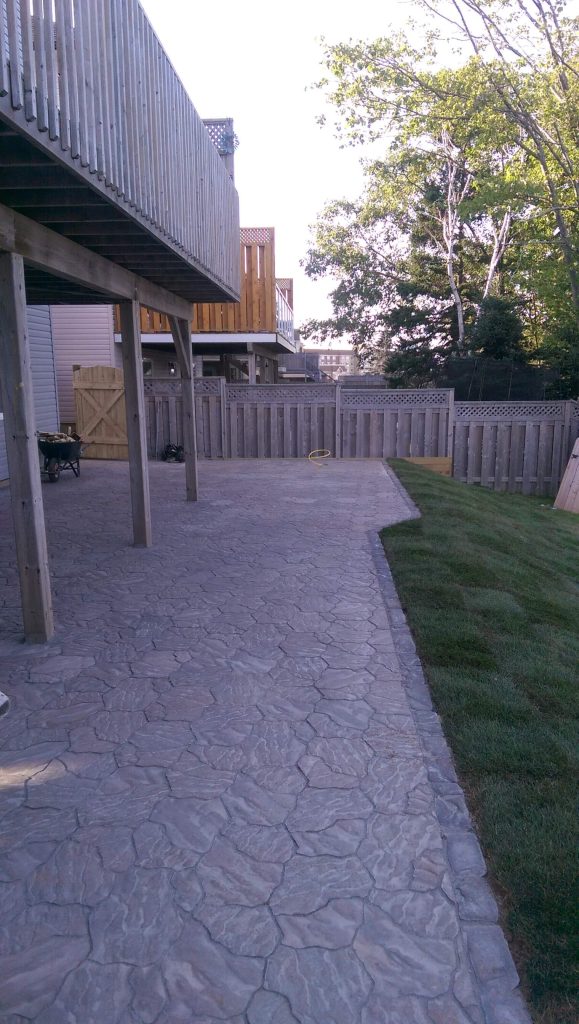 A backyard featuring a concrete stamped patio crafted by Brent's Landscaping, Dartmouth's best. The patio extends from a wooden deck to a grassy area bordered by a wooden fence, with trees in the background. - Brent's Landscaping
