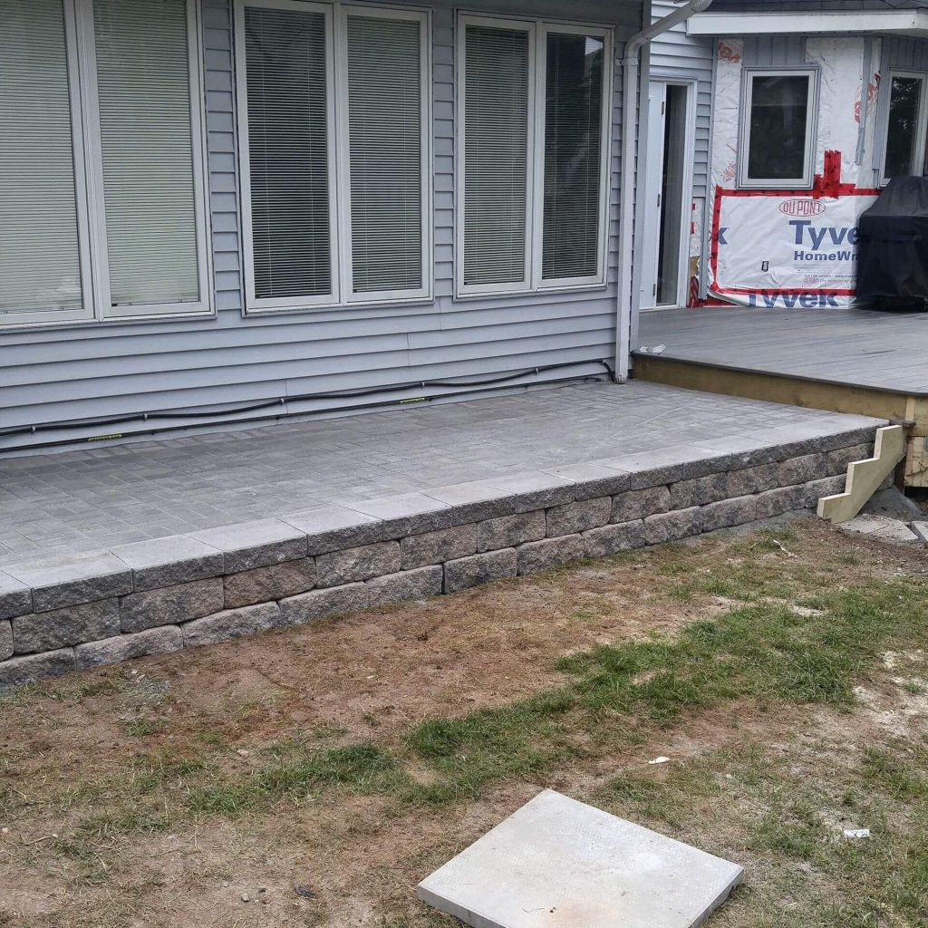 A newly crafted gray stone patio by Brent's Landscaping, Dartmouth's top company, features a low retaining wall. It's set next to a house with large windows and gray siding, with grass and an unfinished area in the foreground. - Brent's Landscaping