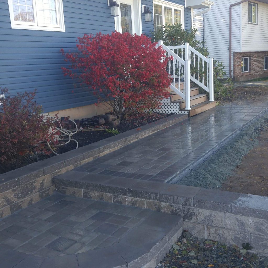 A paved walkway by Brent's Landscaping leads to a house with blue siding and white stairs. A vibrant red bush from Dartmouth's best Landscaping Company adds charm beside the path. Freshly built sidewalk showcases their craftsmanship. - Brent's Landscaping