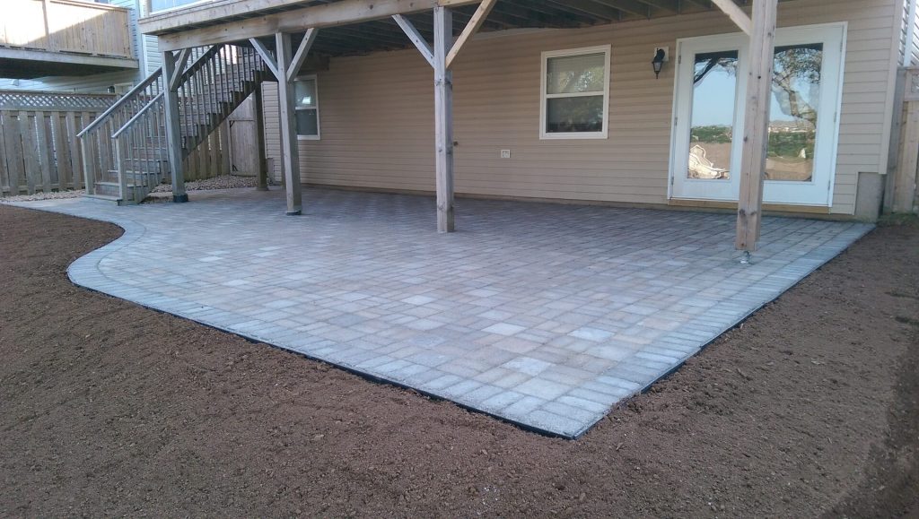 Brent's Landscaping of Dartmouth crafted a stone patio with a geometric pattern under the deck, bordered by rich soil. Beige siding and glass-accented door blend style and nature beautifully. - Brent's Landscaping