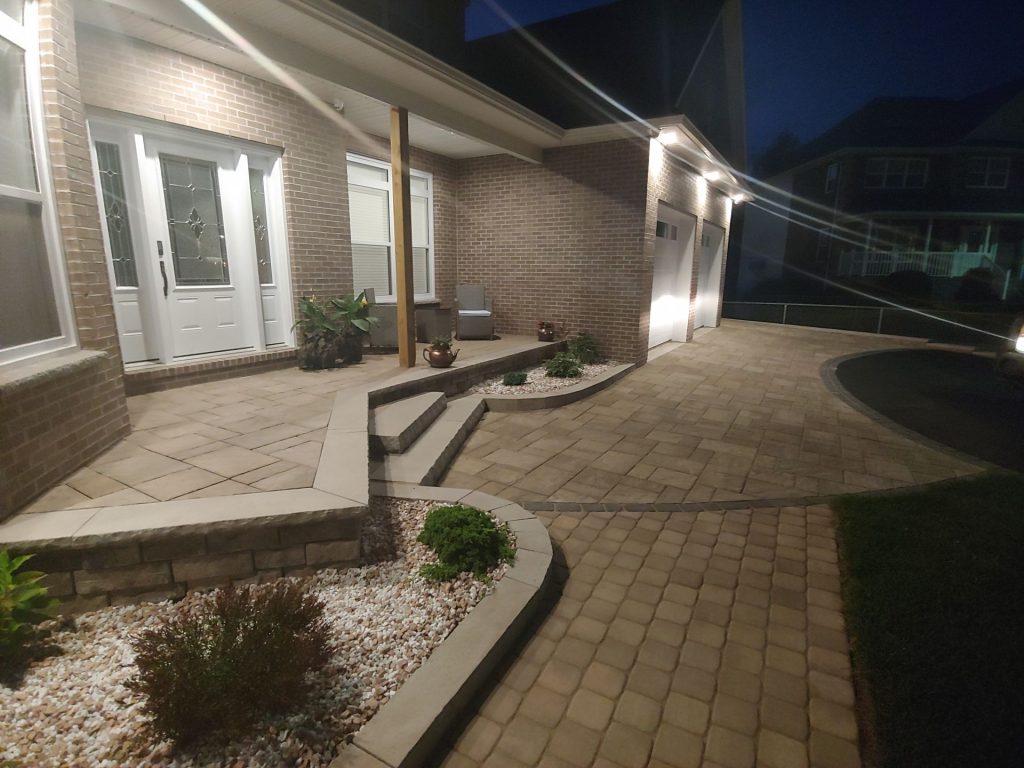 A brick house entrance at night with a well-lit pathway and steps, expertly crafted by Brent's Landscaping, Dartmouth's top landscaping company. The path is bordered by small plants and white pebbles, with lights enhancing the entry doors and garage area for a welcoming look. - Brent's Landscaping