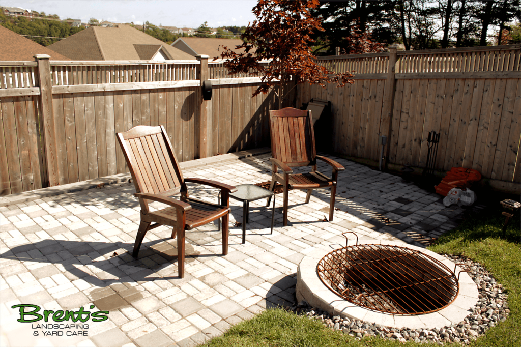 Backyard patio by Brent's Landscaping features two wooden chairs, a small glass table on a stone surface, and a fire pit with grill. Wooden fence and trees enhance this tranquil space, showing why they're Dartmouth’s best. - Brent's Landscaping