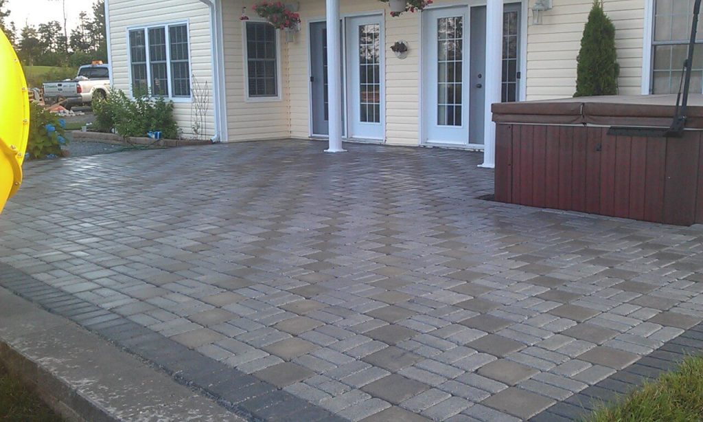 A spacious patio with interlocking stone tiles by Brent's Landscaping extends from a house, featuring a beautifully integrated hot tub surrounded by lush greenery in Dartmouth. - Brent's Landscaping